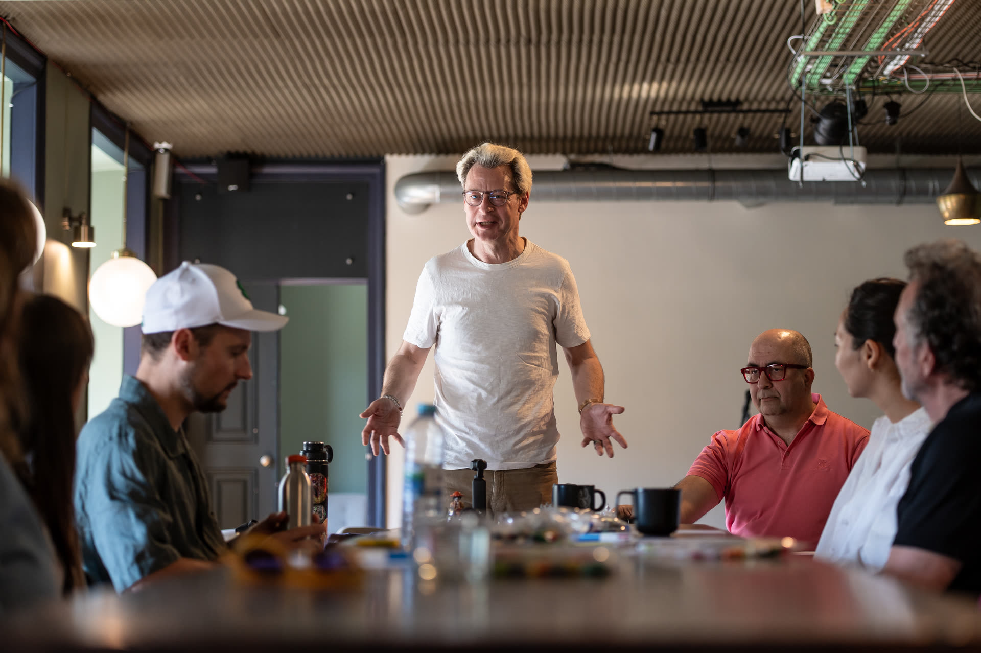 Alexandre Daletchine leading a creativity workshop, standing to address a group seated around a long table scattered with coloured bricks.